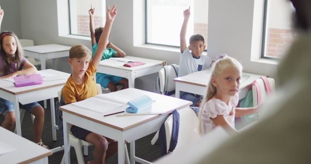 This visual captures elementary school children eagerly raising their hands to answer questions during a class. Each student is seated at their desk in a conducive learning environment. Ideal for use in educational materials, websites for schools or learning resources, and promotional content for educational institutions.