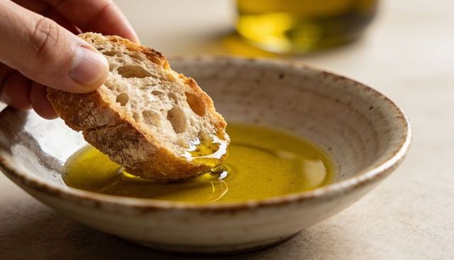 Rustic sourdough dipping into golden olive oil in speckled bowl closeup, mediterranean