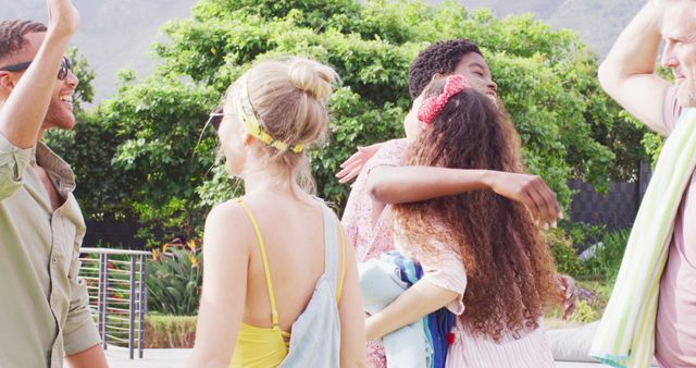 Group of friends hugging and greeting each other outdoors under the sun. The friends are multicultural and dressed in summer attire, implying a relaxed, happy atmosphere. The lush green background and bright light signify a warm, inviting environment. This image could be used for advertisements or promotional materials related to friendship, social gatherings, summer events, vacations, and outdoor activities.