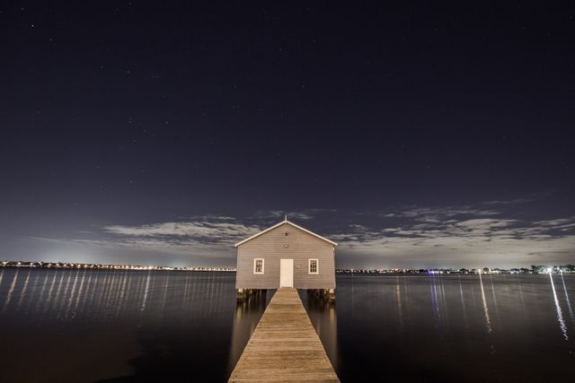 Wooden pier leading to a solitary boathouse on a calm lake at night. Starry sky with reflections on the water creates a serene and tranquil atmosphere. Perfect for illustrating ambiance, peace, solitude, or romantic settings. Suitable for use in travel blogs, inspirational content, relaxation imagery, or wallpapers.