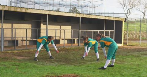 Female softball players in green uniforms warming up near dugout