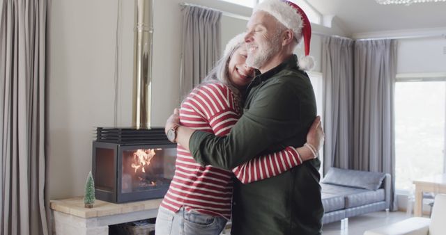 Two seniors wearing Christmas hats are embracing warmly by a lit fireplace. Their home is cozy and decorated for the holiday season, which enhances the cheerful and festive atmosphere. This image can be used for holiday greeting cards, festive advertising campaigns, articles on family and holiday traditions, and social media posts about holiday celebrations and togetherness.