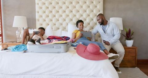 African American family packing suitcase on bed using tablet and phone planning summer trip