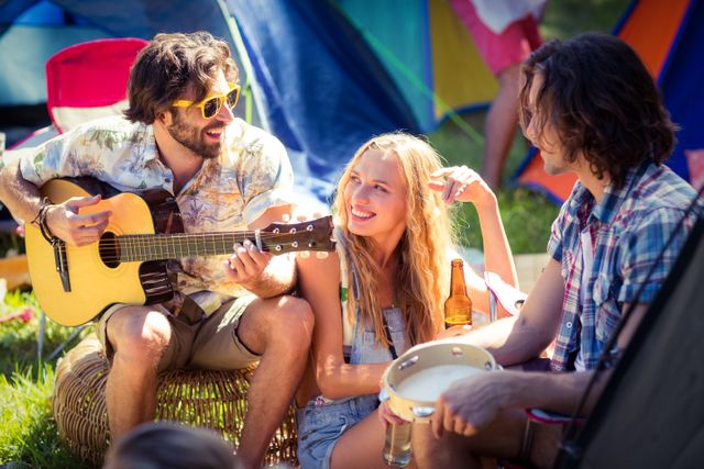 Group of friends enjoying a sunny day at a campsite, playing guitar and tambourine. Ideal for themes related to outdoor activities, summer fun, music festivals, leisure time, and friendship. Perfect for travel blogs, camping gear advertisements, and lifestyle magazines.