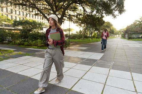 Student walking on sunlit campus pathway holding notebooks, casual college lifestyle