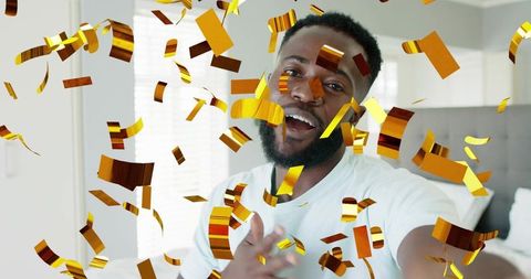 Smiling man reaching toward camera amid swirling gold confetti in bright bedroom celebration