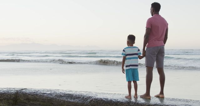 Father standing with his son on the shoreline, both looking at the ocean during sunset. They are holding hands, showcasing a moment of connection and bonding. Ideal for use in family-focused content, advertisements about vacations or insurance, and articles on paternal relationships.