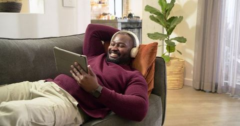 African American man relaxing on sofa with headphones using tablet in modern living room