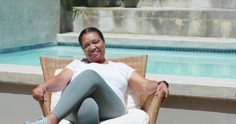 Senior african american woman relaxing poolside in wicker chair smiling in sunny backyard