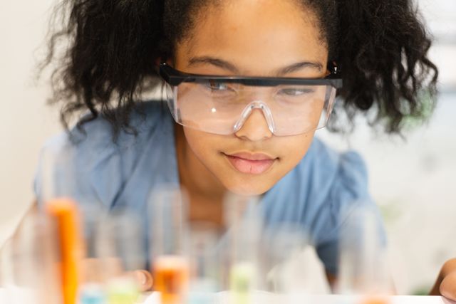 This image depicts a young biracial schoolgirl wearing protective eyewear while observing chemicals in a science lab. Ideal for educational materials, STEM promotion, school science programs, and articles on child education and safety in scientific experiments.