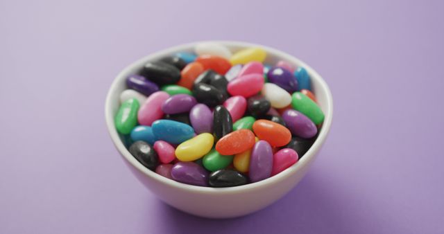 Vibrant and colorful jelly beans in a white bowl are seen on a purple background. Perfect for illustrating themes related to candy, desserts, snacks, and celebration. Ideal for use in advertisements, food blogs, and social media posts to attract attention with its bright and engaging colors.