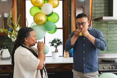 Older asian man and african american woman inflating balloons prepping kitchen party decor