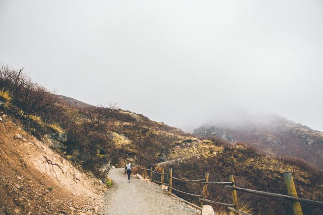 Person hiking on a mountain trail during foggy weather captures the essence of adventure and exploration. Ideal for travel articles, hiking blogs, advertisements for outdoor gear, or motivational content emphasizing journeys and solitude in nature.