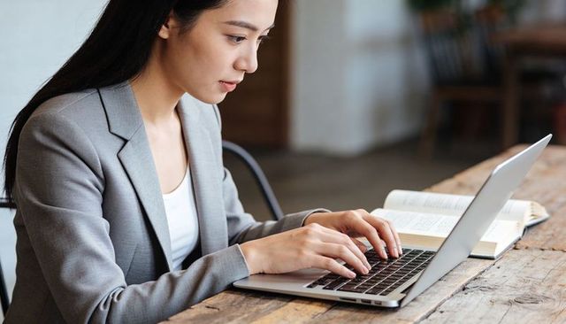 Asian businesswoman typing on laptop in cafe coworking space wearing gray blazer
