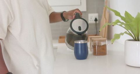 Pouring coffee from electric kettle into blue insulated tumbler on white kitchen counter