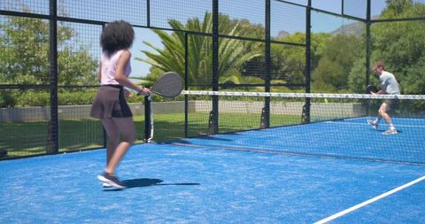 Mixed pair playing padel on bright blue outdoor court, returning shot at net, sunny park