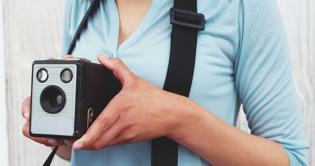 A woman in a light blue shirt holding a vintage camera with a black strap around her neck. Suitably used for themes related to retro photography, hobbies, lifestyle, and vintage technology.
