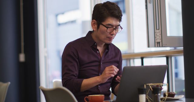 Asian man appears focused while working on laptop in a modern cafe. Ideal for illustrating modern work culture, business environment, freelance workspaces, or urban cafes. Can be used in articles or blogs related to remote work, productivity tips, or technology in professional settings.
