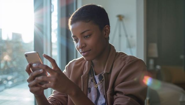 Young black woman scrolling smartphone in sunlit living room wearing brown jacket and gold necklace