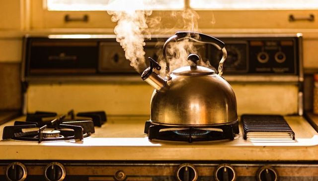 Vintage stainless-steel kettle steaming on gas stove with blue flame and warm sunlight