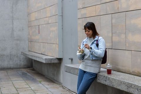 Young asian woman leaning on concrete ledge eating takeout salad in denim jacket