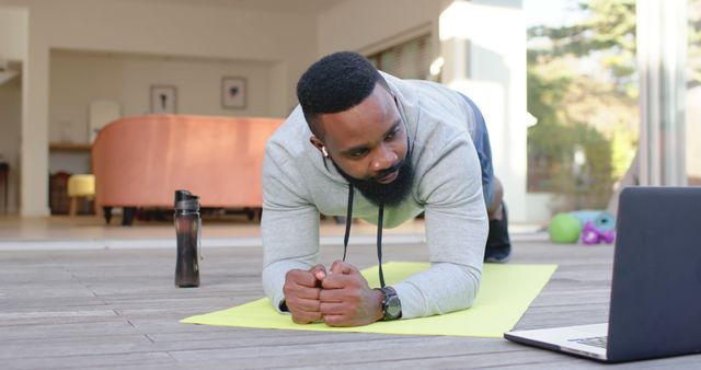 Man performing plank exercise on yellow yoga mat while following a virtual workout on a laptop. He is wearing headphones and engaging in a fitness session at home. Ideal for articles on home fitness routines, virtual exercise classes, and healthy lifestyle.