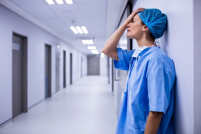 Surgeon in blue scrubs and hairnet leaning against wall in hospital corridor, appearing stressed and exhausted. Ideal for use in articles or presentations about healthcare worker burnout, stress in the medical profession, or the challenges faced by medical staff. Can also be used in healthcare industry reports or mental health awareness campaigns.