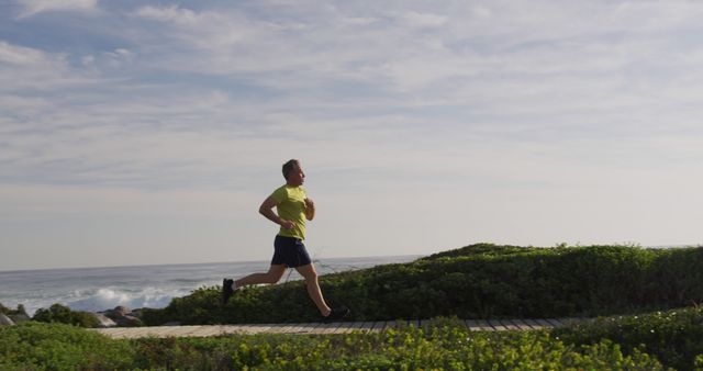 A man jogs along a coastal trail on a sunny day. This photo highlights outdoor exercise and an active lifestyle, featuring lush green foliage and a serene ocean view in the background. Ideal for use in fitness-related promotions, healthy living articles, advertisements for outdoor activities, and health and wellness campaigns.