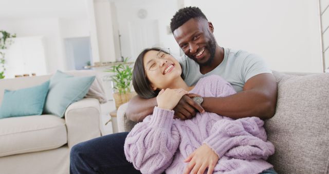 Two people of different ethnicities are cuddling and smiling while relaxing on a couch in a bright living room. This can be used for family, relationship, love, or home comfort-themed content. Ideal for promoting home-related products or services and depicting modern, diverse relationships.