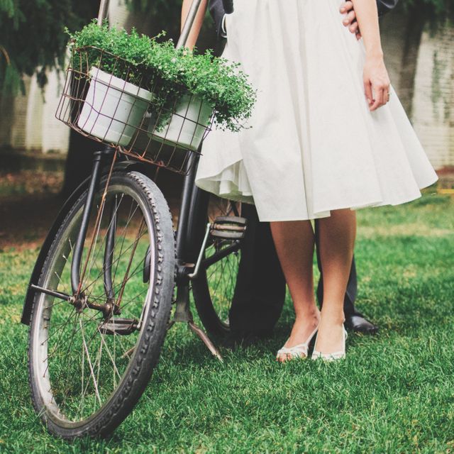 This stock photo showcases a couple's legs standing beside a vintage bicycle with a basket filled with green plants, in a green park or garden. The image exudes a sense of romance, summertime love, and an outdoor lifestyle. It is perfect for use in ads, blogs, or websites focused on nature, vintage lifestyle, love and relationships, outdoor activities, or garden themes.