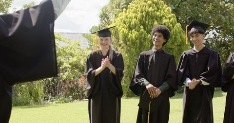 Diverse graduates celebrating outdoors in caps and gowns on sunny campus lawn
