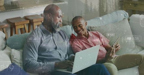 African american father and teen son bonding while using laptop and phone on couch for home tech con