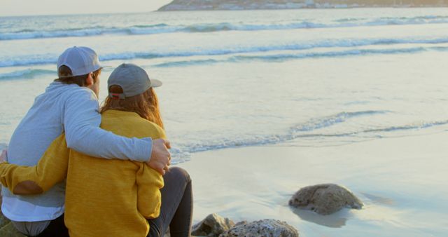 A young Caucasian couple enjoys a serene moment together watching the ocean, with copy space. Their relaxed posture and the tranquil beach setting evoke a sense of peace and companionship.
