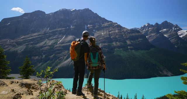 Couple with backpacks hiking on a mountain trail, overlooking a scenic, turquoise lake and majestic mountain peaks on a sunny day. Ideal for use in travel blogs, adventure-themed content, outdoor lifestyle promotions, social media posts, and digital marketing for hiking gear or tourist destinations.