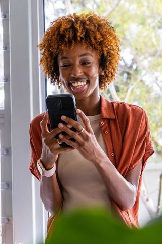 Sunlit african american woman smiling and browsing smartphone near window with houseplant