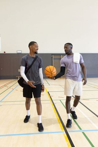 Diverse male friends walking on indoor basketball court passing ball carrying gym bag
