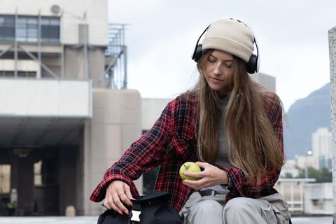 Young woman wearing beanie and headphones sitting on rooftop with backpack and apple