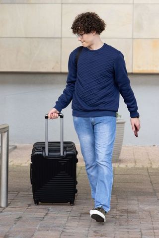 Young man walking with rolling suitcase while checking smartphone in urban street