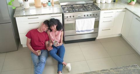 Young diverse couple sharing coffee on tiled kitchen floor leaning together in modern home