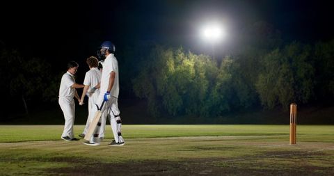 Night cricket team celebrating on pitch under floodlights, diverse players in whites