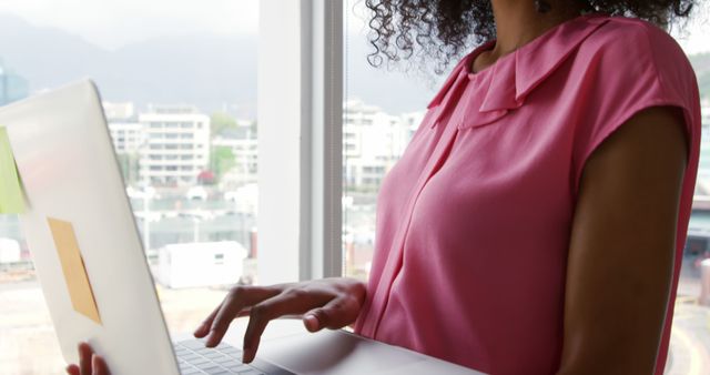 Young businesswoman holding and typing on laptop near large office window. Ideal for use in articles or advertisements about modern work environments, technology in the workplace, remote working, or professional women.