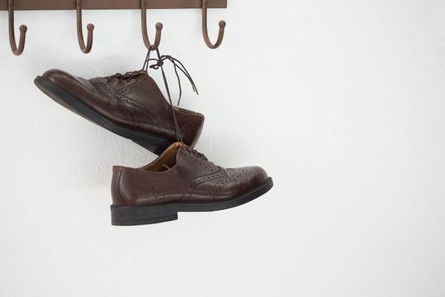 Brown leather shoes hanging on a wall hook against a white wall. Ideal for concepts related to organization, minimalist home decor, and storage solutions. Suitable for use in articles, blogs, and advertisements focusing on interior design, tidiness, and footwear storage ideas.