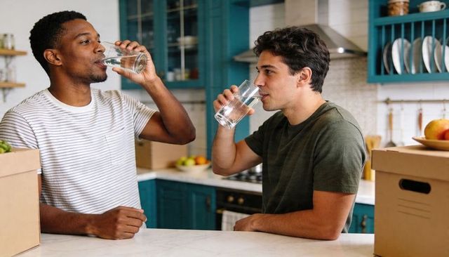 Two friends drinking water at kitchen island with moving boxes and teal cabinets