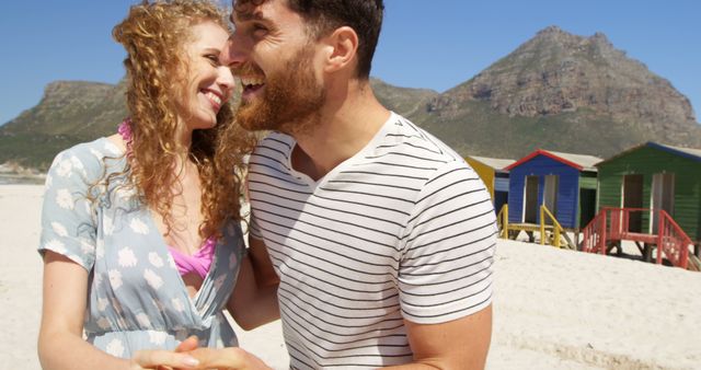 A young Caucasian couple is sharing a joyful moment on a sunny beach with colorful beach huts in the background, with copy space. Their smiles and close proximity suggest a romantic connection as they enjoy the beautiful coastal setting.