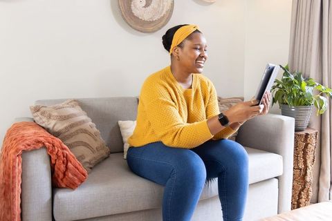 Smiling african american woman using tablet on cozy gray sofa at home, mustard sweater