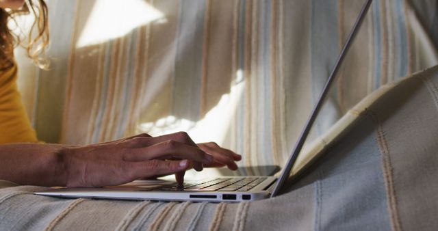 Person typing on laptop while sitting or lying down on couch with striped blanket in a cozy home setting. Can be used for concepts related to remote work, freelance, online study, casual computing at home, technology use in relaxed environments, digital tasks.