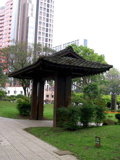 Traditional Japanese gate stands among modern high-rise buildings in an urban park. Provides contrast between cultural heritage and city development, suitable for themes involving cultural integration, architecture, green spaces in cities, or Asian influences in urban design.