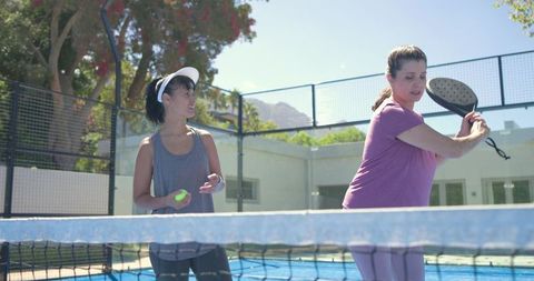 Two women playing pickleball on sunny outdoor court, one swinging paddle while partner watches