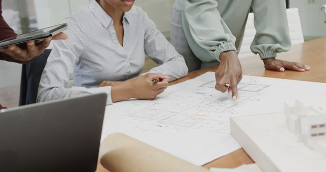 Group of architects examining architectural blueprints in a modern office environment. The photo captures an African American woman taking notes while collaborating with others. Ideal for use in business, architecture firm websites, team collaboration articles, and project planning presentations.