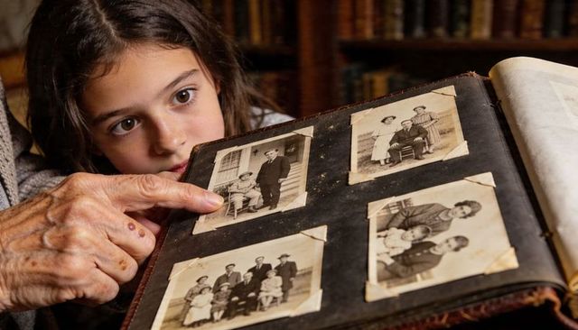 Grandparent pointing through vintage family album, grandchild discovering generations of memories